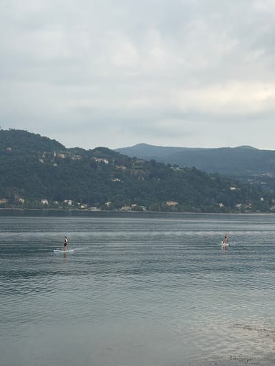 The image depicts a body of water with a wooded, hilly landscape in the background. The sky is overcast. Two individuals are visible on paddleboards in the water. The person on the left is standing on a paddleboard, facing towards the right. The person on the right is also standing on a paddleboard, with another individual behind them, appearing to be sitting. The water exhibits rippling patterns. The background shows a densely forested hillside dotted with buildings, suggestive of a town or village. The scene is likely a lake or a large river, with a backdrop of the Italian landscape, possibly near Angera, Italy, known for its lakeside setting and hillside villages.