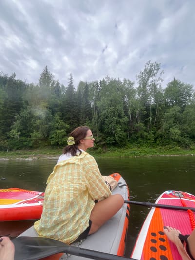 The image depicts two individuals on paddleboards on a body of water, with a dense forest lining the opposite bank. The sky is overcast with clouds. In the foreground, a person is seated on a paddleboard, facing to the right and looking towards the forest. They are wearing a yellow plaid shirt and sunglasses. Another paddleboard is partially visible to the right, with a person's feet on it. The paddleboard in the foreground has a red and white design with orange accents. The water appears calm and green. The trees in the background are a mix of evergreen and deciduous species, with lush green foliage. A paddle is visible in the lower left corner of the frame. The location is identified as the city of Manskii, Russia.