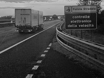 A black and white, medium-shot, eye-level photograph captures a highway scene at dusk near Somaglia, Italy. In the foreground, the asphalt road displays dashed white lane markings. A metal guardrail is positioned to the right, with a dark sign above it. The sign has two sections. The top section, rectangular and white, contains a silhouette of a person in uniform and the text "Polizia Stradale". The bottom section, a dark rectangle, bears white text that reads "controllo elettronico della velocità".
In the mid-ground, a white semi-trailer truck, bearing the "SCHENKER" logo on its side, travels in the left lane. The truck has illuminated headlights and taillights. Further down the highway, more vehicles and an overpass are visible in the hazy distance. The sky above is overcast with clouds. The overall impression is that of a functional, albeit somber, highway environment under low light conditions.