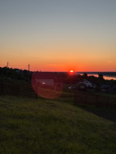 The image captures a rural or suburban landscape in Зеленец, Russia, at either sunrise or sunset. The sky displays a gradient from light blue-grey at the zenith to a vibrant orange-red at the horizon, where a distinct, bright orange-red solar disk is partially visible above distant landforms. A singular small avian entity is discernible in the upper left sky. A significant red-orange circular lens flare, originating from the sun, extends across the lower right portion of the image, impacting a green grassy slope and a dark wooden multi-rail fence. The foreground consists of a densely vegetated green grassy slope, with darker, possibly tilled earth visible further up. The midground contains several residential houses with varying architectural designs and dark roofs, interspersed among dense deciduous trees. Utility poles with overhead power lines are also present. A broad body of water, reflecting the intense orange sunlight, horizontally spans the background, with its opposite bank lined by dark tree cover. No human or other animal subjects are present.