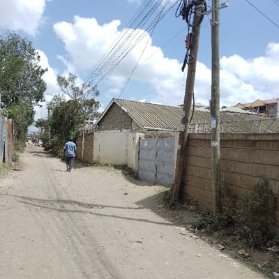 An outdoor scene in Ongata Rongai ward, Kenya, depicts a dirt road under a partially cloudy blue sky. A single male subject is walking away from the camera, centered on the unpaved road. He wears a light-colored, possibly plaid, long-sleeved shirt and dark trousers. Ahead of him, in the distance, indistinct figures and structures are visible along the road, with at least one light-colored vehicle present.

To the right, a light-colored concrete block wall, approximately 2 meters high, runs parallel to the road, topped with a security barrier resembling barbed or razor wire. A grey, double-leaf metal gate is set into this wall. Behind the wall, a building with a pitched roof covered in light-colored corrugated sheets is visible. Further right, two wooden utility poles stand near the wall. The pole closer to the camera is slightly angled, laden with numerous black electrical or communication wires and attached equipment. The second pole is straighter and less burdened. Multiple overhead wires span across the frame from right to left.

To the left of the road, a corrugated metal fence borders the scene, with dense green foliage and trees growing along it and extending further into the background. Sparse vegetation, including small bushes and weeds, grows along the edges of the dirt road, particularly on the right side near the wall. The road surface exhibits some tire tracks and minor irregularities.