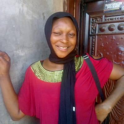 A close-up image depicts a single adult female in Ibreda, Nigeria, standing before a background composed of a light grey, textured wall on the left and a dark brown, ornately carved wooden door on the right. The female, possessing dark skin, wears a black headscarf that covers her hair. She is dressed in a red, loose-fitting top with short, wide sleeves, distinguished by multiple rows of golden, possibly beaded or sequined, embellishments encircling the wide neckline. A black strap, indicative of a shoulder bag, traverses her left shoulder. Her right arm is slightly raised and bent at the elbow, with the hand open, while her left hand is positioned on her hip. She faces forward, directly engaging the viewer with a broad smile. Affixed to the upper right portion of the wooden door is a white rectangular sticker or poster, clearly displaying the black text "MY YEAR OF TRANSFORMATION & OPEN HEAVEN."