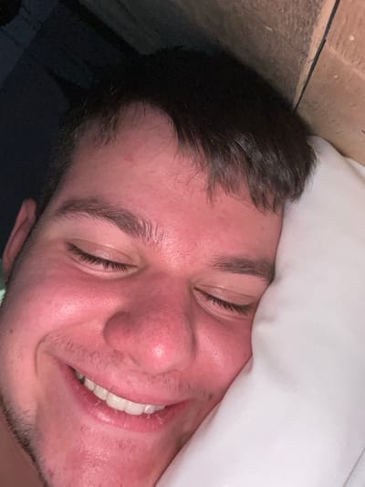 A close-up image shows a man lying in bed, appearing relaxed or asleep with a slight smile. He is positioned on his side, with a white pillow partially covering his forehead. His eyes are closed, and his dark hair is tousled. The man has fair skin, dark eyebrows, and stubble. To the upper left, there is a wooden surface. The location, Pamporovo, Bulgaria, is not discernable from the image.