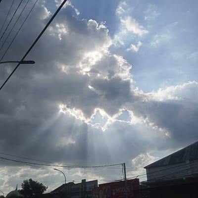 The image depicts a street scene in Asembagus, Indonesia, under a dramatic sky. Thick, dark clouds dominate the upper portion of the frame, with the sun's rays breaking through in a powerful crepuscular display. The sunbeams emanate from a break in the clouds, casting ethereal lines of light downwards. In the foreground, power lines crisscross the sky, obscuring parts of the view. A street lamp is visible on the left side of the image. Below the sky, the rooftops of buildings are visible, one with a corrugated metal roof. A green domed structure, possibly a mosque, is partially visible on the far left. Trees and other buildings, including a storefront with a red sign that says "MIXUE," are also present. The overall impression is one of a dynamic and atmospheric weather event occurring within a semi-urban environment. There are no people or animals clearly visible in the image.