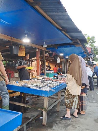 The image depicts an outdoor market scene in Cottarum Tunong, Indonesia. Several individuals are present, some appearing to be vendors and others customers. In the foreground, a person in a checkered shirt and dark apron is holding a yellow object, possibly a bottle or container. To the right, a stall displays a large quantity of fish, laid out on a blue surface. The market appears to be covered by a corrugated metal roof and blue tarpaulins, supported by wooden structures.
Several people are seen in the background, some wearing traditional Indonesian clothing, including sarongs and headscarves. One individual is wearing a brown hijab and a light-colored garment. The overall setting suggests a bustling marketplace, likely in the morning or afternoon given the daylight. The ground appears to be paved or a dirt surface. There are no explicit cues about the weather, but the lighting suggests it is daytime.
Visible text includes "SAKURA" on the blue tarpaulin, likely a brand name. The scene conveys a sense of commerce and daily life in a local Indonesian community.