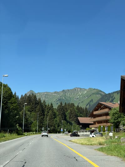 This is a vertical, slightly angled, full shot of a scenic mountainous area with a road running through it. The scene depicts a clear, bright blue sky with a very small crescent moon visible high up on the left. The right side of the frame is dominated by a lush green mountainside with patches of snow near the peak. In the foreground on the right, a paved road with a yellow lane marker curves away.
Along the road, several buildings with traditional chalet-style architecture, featuring wooden balconies and brick accents, are visible. Some of these buildings appear to be residences or hotels. There are also numerous pine trees lining the road and clinging to the lower slopes of the mountain. Several streetlights are positioned along the side of the road.
Two cars are visible on the road: a white car further up the road and a black car closer to the foreground, both parked. A few traffic signs and lampposts are also present. The overall setting suggests a sunny day in a mountainous, possibly rural or resort, location. The time of day appears to be daytime, likely late morning or early afternoon, given the bright sunlight and clear sky. There is no discernible text in the image.