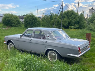 A gray GAZ-24 Volga sedan is parked in tall, green grass. The car has whitewall tires and chrome accents. In the background are green trees, power lines, a utility pole, and a water tower. The scene suggests a rural or suburban area of Troitsk, Russia. A rusty metal barrel is visible near the rear of the car.