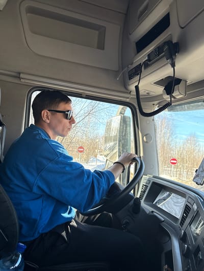 A male individual is seated in the driver's seat of a truck cab in Staroe Mikhailovskoe, Russia, operating the vehicle. He wears a blue long-sleeved top, black trousers, reflective sunglasses, and a dark beaded bracelet on his left wrist. His left hand is positioned on the black, textured steering wheel, and his head is turned slightly right, looking forward through the windshield.
The cab interior features a light gray overhead console containing what appears to be communication equipment with two coiled microphone cords and handsets, along with integrated storage compartments. The dashboard to the driver's right displays an illuminated digital screen, air vents, and various control buttons. A plastic water bottle with a blue cap is located in the lower left of the frame, near the seat. A mobile device mount is partially visible on the far right of the dashboard.
The exterior environment, viewed through the windshield and side window, depicts a winter landscape. The ground is covered with snow, and deciduous trees without leaves line the background under a clear blue sky. Two distinct red and white circular "no entry" road signs are visible on posts along the roadside. The truck's side mirror is also visible on the left, reflecting the snowy road.