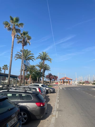 The image depicts a street scene in Canet d'en Berenguer, Spain, featuring a row of parked cars on the left side of the road, facing away from the viewer. Palm trees are prominent throughout the scene, lining the street and the background. To the left, a restaurant with outdoor seating is visible, with tables and chairs set up. Further down the street, a small gazebo with an orange tiled roof stands on a pedestrian island. Beyond the gazebo, a marina with numerous sailboats docked is visible. The sky is clear and blue with some wispy clouds and a contrail from an aircraft. The ground is asphalt with painted road markings. The cars appear to be parked legally along the curb. No people are clearly depicted engaging in any specific actions. The overall atmosphere is one of a sunny day in a coastal town.