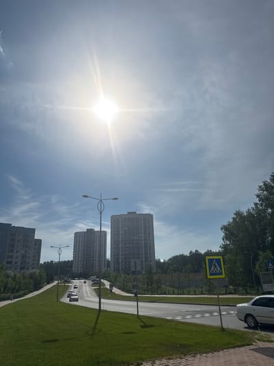 A sun-drenched scene captures a modern cityscape in Koltsovo, Russia, under a bright blue sky dotted with wispy clouds. The sun, a brilliant starburst, dominates the upper portion of the frame, casting a strong glare. In the foreground, a neatly mown grassy hill slopes down towards a paved road. The road curves gently upwards, with several cars driving on it.
Tall, multi-story apartment buildings line the sides of the road, their modern architecture standing out against the natural backdrop of green trees. Streetlights with distinctive oval designs are strategically placed along the road, illuminating the urban environment. A yellow pedestrian crossing sign with a silhouette of a person walking is prominently visible on the right side of the road, indicating a crosswalk. A white car is parked near the pedestrian crossing. The presence of these elements suggests a well-developed residential area with functional urban infrastructure. The overall impression is one of a peaceful, everyday scene in a contemporary Russian city.