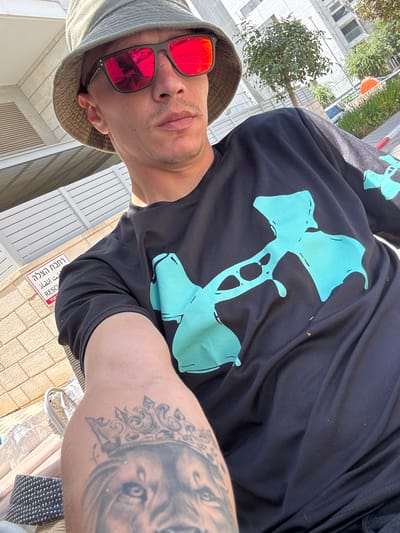 A person is captured in a close-up, low-angle selfie. The subject is wearing a dark t-shirt with a prominent light blue graphic design. They are also wearing a light-colored bucket hat and red-tinted sunglasses, which reflect the surrounding environment. The background reveals an outdoor setting with some greenery, a building, and a fence. A sign with text in Hebrew is visible on the ground, reading "כניסה אסורה" and "RESC". The lighting suggests it is daytime, possibly sunny. The overall impression is a casual, personal snapshot taken outdoors.