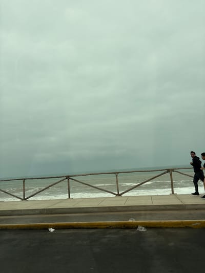 The image shows a portion of a walkway or promenade overlooking the ocean. Two individuals, appearing to be young men dressed in dark clothing, are visible in the upper right portion of the frame, standing near a wooden railing. The railing is constructed from rustic-looking wooden poles and planks, forming a triangular pattern. Beyond the railing, the ocean stretches out with visible waves. The sky is overcast and a pale gray, suggesting a cloudy or hazy day. The ground in the foreground appears to be asphalt with a yellow line marking its edge. A couple of pieces of white plastic litter are visible on the asphalt near the yellow line. The scene is oriented vertically, with the horizon line running horizontally across the middle of the image. The overall impression is a simple, everyday scene by the sea on a rather dull day. There is no visible text in the image.