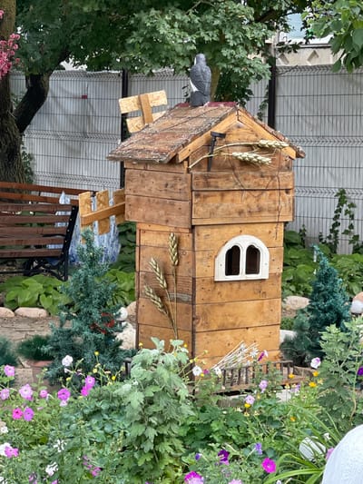 In Budyonnovsk, Russia, a whimsical wooden birdhouse, designed to resemble a multi-story structure with a peaked roof and an arched window, is nestled amongst lush greenery and vibrant flowers. A ceramic bird figurine perches atop the roof, surveying the scene. The birdhouse is adorned with decorative wheat stalks and a small solar-powered light. In the background, a metal fence separates the garden from a paved area, and a wooden bench is partially visible. The garden itself is a riot of color, with pink and purple petunias, yellow buttercups, and various other blooming plants creating a picturesque and lively atmosphere.