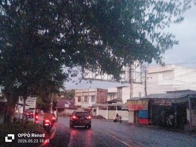 The image depicts a street scene in Peub Lueng Nibong, Indonesia on what appears to be a rainy or overcast evening, as indicated by the wet road surface and dim lighting. The image has been taken by an OPPO Reno8 phone, as indicated by the watermark. The time of the image capture is recorded as 18:43 on May 23, 2026. A tree with dense foliage occupies a significant portion of the upper left and center of the frame, partially obscuring the background. The road is visible in the lower portion of the image, with vehicles present. A black car with illuminated tail lights is in the center, facing away from the camera, with another two motorbikes either in front or alongside it, also with illuminated tail lights. A fourth motorbike can be seen on the right of the road. Buildings and structures are visible along the roadside, including what appears to be a small shop or stall with a colorful facade on the right. A large billboard structure can be seen in the background. Utility poles and overhead wires are also present. The scene suggests a residential or commercial area during dusk or evening.