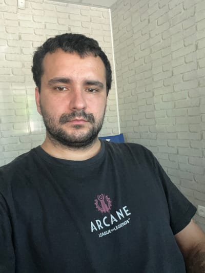 A man, with dark hair and a beard, is shown from the chest up, facing the camera at a slightly upward angle. He is wearing a black t-shirt with the word "ARCANE" printed in large white letters diagonally across the chest. Below "ARCANE," the words "LEAGUE OF LEGENDS" are printed in smaller white text. A circular, stylized pink graphic is visible to the left of the text. The background is a light-colored, brick-patterned wall. The lighting suggests it is daytime. The man's expression is neutral, and he is looking directly at the camera. The orientation of the photo is such that the man appears to be standing or sitting upright, with the background wall behind him. The image appears to be taken indoors.