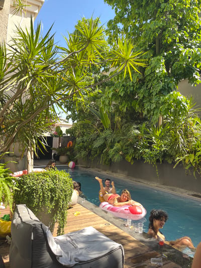 An outdoor scene in Binyamina - Givat Ada, Israel, depicts a group of individuals engaged in leisure activities around a narrow, rectangular swimming pool. Six people are visible. In the immediate foreground, a dark grey bean bag chair with a light-colored towel rests on wooden decking. Adjacent to it, a female with dark, curly hair, wearing a black bikini top, is partially submerged in the pool, smiling and looking right. Several clear plastic cups and one glass are on the wooden edge of the pool near her.
Towards the center, a female wearing a red bikini lies face-up on a pink inflatable float, smiling at the camera. Behind her on the same float, a male with short dark hair and a beard is partially visible, lying on his stomach with legs raised. To the left, a female with light hair and sunglasses is partially obscured by foliage, in or near the water. Further in the background, another individual is partially visible in the pool near a colorful beach ball. A red foam noodle float is near the far left edge of the pool.
The pool area is surrounded by dense, lush green vegetation, including various palm-like plants, large-leafed plants, and hanging foliage. In the background, a dark slatted fence or wall is visible, with a large potted plant and an orange life ring hanging on the fence. Portions of light-colored building structures are visible in the upper left. The scene is illuminated by bright daylight under a clear blue sky.