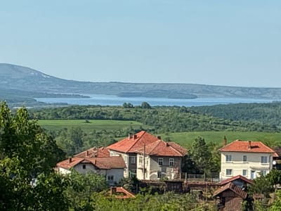 The image depicts a rural scene in Bokilovtsi, Bulgaria, featuring houses with red tile roofs nestled amidst green trees and foliage. In the background, a body of water is visible, with distant hills or mountains beyond. The sky is clear and light blue. The foreground is dominated by trees, partially obscuring the view of the houses. The houses appear to be located on a slightly elevated terrain, giving them a higher vantage point over the surrounding landscape.