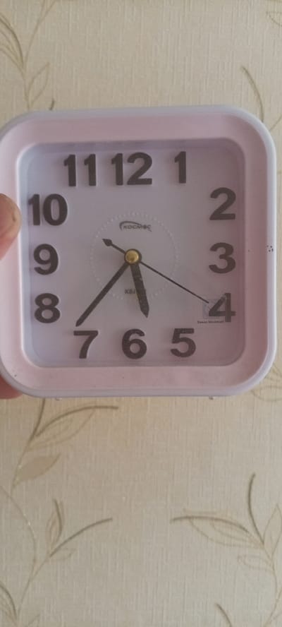 A close-up, eye-level shot shows a square, pink analog alarm clock against a lightly textured, beige wallpaper with a faint floral pattern. The clock face is white with large, black Arabic numerals. The hour hand points to the number 4, and the minute hand points to the number 7, indicating the time is approximately 4:35. The second hand is visible and points slightly past the 2. A circular logo with Cyrillic text "КОСМОС" (KOSMOS) is present on the clock face, along with "Sweep Movement" text near the number 4. The clock is placed on a surface that is not visible in the frame. The wallpaper suggests an indoor setting. This image does not provide information about a specific location in the city of Zhigulyovsk, Russia, as it only shows a close-up of a clock.