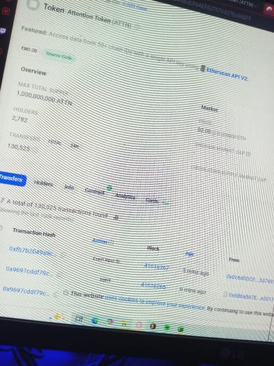 The image displays a computer monitor positioned in a dark environment with a subtle blue ambient light visible at the bottom. The monitor, bearing an "LG" logo on its lower bezel, exhibits horizontal scan lines, indicating a photograph taken of a digital screen. The screen shows a webpage titled "Token Attention Token (ATTN)," likely a cryptocurrency or blockchain explorer interface, located in Tiruchirappalli, India.

The webpage provides an "Overview" of the ATTN token, listing a "MAX TOTAL SUPPLY" of 1,000,000,000 ATTN, 2,782 "HOLDERS," and 130,525 "TRANSFERS TOTAL." A "Market" section indicates a "PRICE" of "$0.00 @ 0.000000 ETH." Navigation tabs include "Transfers," "Holders," "Info," "Contract," "Analytics," and "Cards," with "Transfers" highlighted. Below, a table details recent transactions, showing "Transaction Hash," "Action," "Block," "Age," and "From" fields. Two visible transactions include hash "0xfb7b204939c..." with "Action: Execut Input Sr," "Block: 41516367," "Age: 5 mins ago," and a "From" address "0x0c6d0DCE...3d7491," and hash "0x9697cddf79c..." with "Action: Inject," "Block: 41516265," "Age: 9 mins ago," and a "From" address "0x6B6a567E...e30111." A cookie consent banner partially visible at the bottom of the screen reads, "This website uses cookies to improve your experience. By continuing to use this web..." No individuals are present in the image.