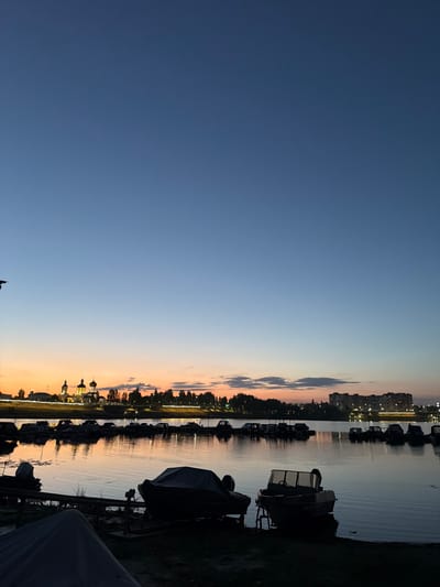 This is a photograph of the Volga River waterfront in Syzran, Russia, at dusk. The sky is a gradient of deep blue to pale orange, indicating the sun has recently set. On the far bank, a cluster of buildings, including a church with golden domes, is illuminated by artificial lights. Across the river, a marina is filled with numerous boats docked and covered. Some boats are also visible on the near bank, with one small motorboat on a trailer in the foreground, partially covered. The water reflects the sky and the lights from the distant shore. The overall atmosphere is calm and serene.