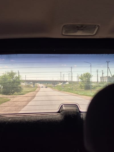 The image, taken from inside a vehicle looking out the rear window, captures a road leading towards a bridge under a clear sky with scattered clouds. The road is paved and appears to be a two-lane highway. To the left of the road, there is a grassy area with trees and bushes. On the right side of the road, there are utility poles with power lines and a few buildings in the distance. There are several cars on the road, with two visible in the foreground and a few more further down the road. The interior of the vehicle is visible at the bottom of the image, with the headliner and a rear-view mirror housing at the top. The location appears to be rural or semi-rural, possibly on the outskirts of a town or village. The scene is consistent with a view from a vehicle traveling on a highway in Kazakhstan, possibly in the vicinity of Новоишим ауылдық округі.