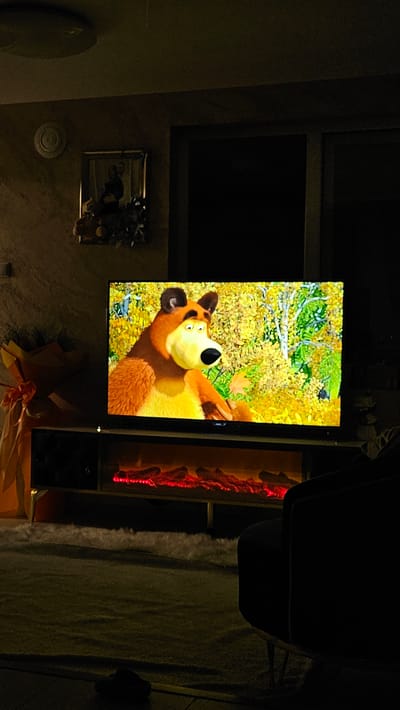 The image depicts an interior scene, likely a living room, in Iasenovets, Bulgaria, characterized by low lighting conditions suggesting evening or nighttime. A flat-screen television, positioned centrally, displays a frame from the animated series "Masha and the Bear," featuring the brown bear character with a surprised expression against an autumn forest background with yellow and green foliage. The television rests on a dark entertainment unit, which incorporates an electric fireplace with a simulated red glow of burning logs in its lower section. To the left of the television, a large, vertically oriented object wrapped in orange or yellow paper with ribbons, possibly a gift or bouquet, stands on the floor. Above this object, a framed picture hangs on a textured, light-colored wall, depicting two indistinct figures. A circular, white wall ornament is visible further to the left of the picture. To the right of the television, a dark-colored armchair is partially visible. The floor is covered with a dark carpet, with a lighter, possibly shaggy, rug segment in front of the entertainment unit. A dark, rectangular shape, likely a window or door, is visible in the upper right background. A ceiling light fixture is dimly visible at the top center. No individuals are present in the observable scene.