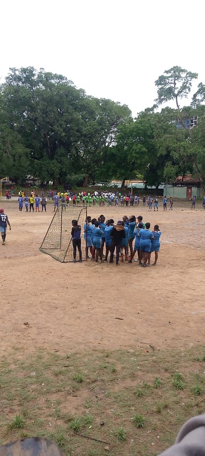 The image depicts an outdoor sports field, consistent with a location in Tudor ward, Kenya, under an overcast sky. The ground consists of exposed reddish-brown earth with sparse patches of green grass and scattered small debris.
In the midground, a portable soccer goal with a dark net stands to the left. A group of approximately 15-20 individuals, predominantly female, wearing light blue athletic jerseys and shorts, are huddled tightly together near this goal, with one central figure in a dark jacket appearing to address them. Another individual in dark attire stands near the goal. To the far left, a person wearing a blue jersey with the number "12" is visible walking away from the camera.
Further in the background, multiple groups of people are scattered across the field. Some are aligned in formations, wearing uniforms of varying colors, including blue/yellow and light green/white. A basketball hoop structure is partially visible on the far left.
The background features a dense treeline of large, mature trees with green foliage. Behind these trees on the right, the concrete framework of a multi-story building is partially visible, along with smaller structures exhibiting reddish roofs.