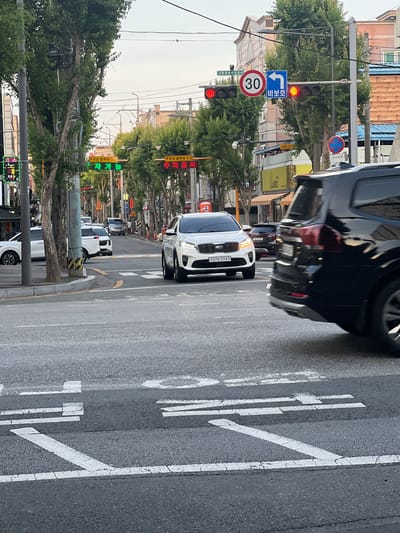 The image shows a street scene in Gwangju, South Korea. In the foreground, a white Kia Sorento is stopped at a crosswalk, its headlights illuminated. To the right, the rear of a black SUV is partially visible, also in motion. In the background, several other vehicles, including white SUVs and cars, are navigating the street. Traffic lights display red and yellow signals, indicating a stop or caution. Road signs are visible, including a "30" speed limit sign and a blue sign with a left-pointing arrow and Korean text that likely indicates a turn. Trees line the street, casting shadows. Buildings, including some with light-colored facades and orange accents, are visible in the distance. The sky is overcast. The road surface is asphalt with visible white lane markings and pedestrian crossing lines. The overall impression is that of a typical urban street intersection during daylight hours.