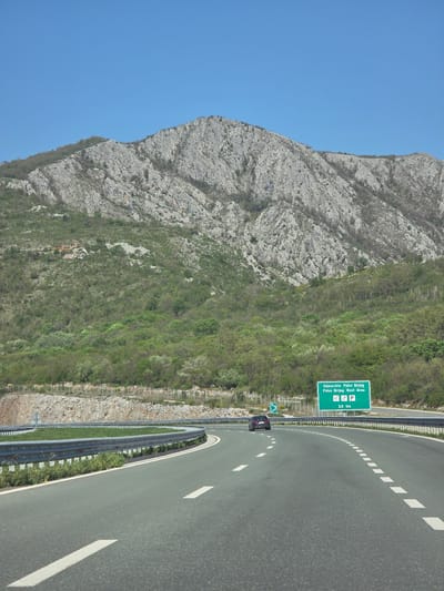 The image depicts a highway in Pelev Brijeg, Montenegro, adjacent to a mountain. The road curves slightly, with white lane markings. A sign indicates a rest area "Odmorište Pelev Brijeg" with parking and restroom facilities. A single car is visible on the road. A crash barrier separates the road from the mountainous terrain. The mountain is rocky and covered in sparse vegetation. The sky is clear and blue.