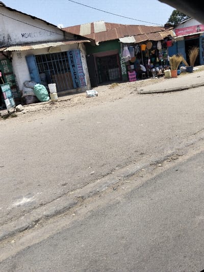 The image depicts a street scene in Mkomani ward, Kenya, during daylight hours under a clear sky. The foreground and mid-ground are dominated by a worn asphalt road with visible cracks and patches. On the right side, the road appears to slope slightly upwards.
Along the left and center background, a series of single-story commercial buildings line the street. From left to right:
1. A building with a white-painted facade and a corrugated iron roof, featuring a light blue security-barred window. A vertical sign on the wall reads "CHEMICALS." Outside this building, green and white/grey sacks, along with white plastic buckets, are piled on the dirt verge. A green advertising board is also partially visible on the far left.
2. Adjacent to the first building, a green-painted storefront with a brown corrugated iron roof is visible. This shop displays various household items, including orange, yellow, and red plastic buckets, basins, and what appear to be textiles or clothing hanging outside. Brooms are arranged vertically near the entrance. One individual is discernible sitting inside or directly in front of this shop, partially obscured by merchandise.
3. Further right, a partially visible blue-painted storefront with a corrugated iron roof bears a sign that reads "SEDO AGENCIES." More brooms are positioned outside this establishment.
A single dark utility wire traverses the upper portion of the sky. The overall environment appears to be a busy commercial area. No discernible interactions between people are visible.