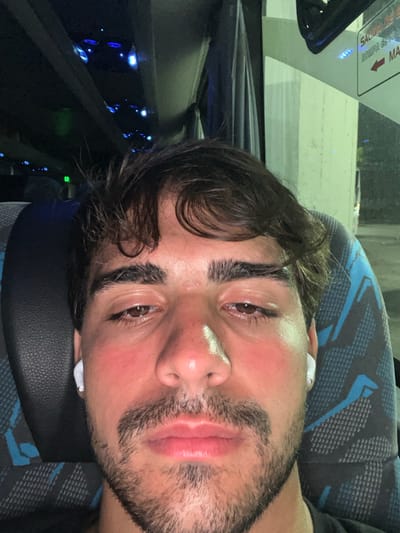 A man with brown hair, stubble, and flushed skin rests on a patterned bus seat. He wears wireless earbuds. The interior of the bus is visible, with a window showing a white structure outside and illuminated blue lights within the vehicle. The image is rotated, giving a tilted perspective of the man's face and surroundings.