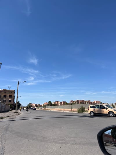 The scene shows a street in Tassoultante, Morocco, with a clear blue sky dotted with wispy clouds. A beige taxi cab is parked on the right side of the street. A tan brick wall runs along the roadside, interspersed with buildings of similar construction. A street sign, partially obscured, is mounted on a post along the wall. The road surface is paved. A building and several utility poles are visible near the bottom of the frame. There are motorbikes and people further down the road. A car's side mirror is visible at the very right.