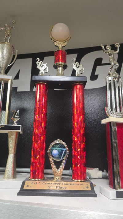 The image shows a trophy display on a shelf in what appears to be an indoor setting. The trophy in the center is a large, elaborate piece with two red, faceted columns topped with a gold decorative element featuring the numbers "2013" on either side. Above this is a gold globe with a volleyball on top. Below the red columns is a black base with a gold plaque that reads "EvCC Crossover Tournament 2ND Place". To the left of the central trophy, there is another trophy with a gold figure of an athlete holding a ball and a large silver cup. To the far left, a tall, thin trophy with a gold base and a silver column is visible. To the right of the central trophy, a tall, red and gold trophy is partially visible. The background is a black wall with a large white graphic, possibly a logo or lettering. The shelf is white and appears to be made of laminate. No people are visible in the image. The location is identified as the city of Bremerton, United States of America.