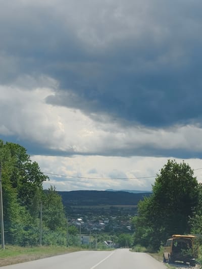 The image shows a gray asphalt road leading towards a distant village nestled in a valley. The sky is heavily clouded with dark, ominous storm clouds dominating the upper half of the frame, interspersed with patches of lighter clouds. Lush green trees line both sides of the road, with a dense forest on the left and scattered trees on the right. A small, yellow van with its rear hatch open is parked on the side of the road on the right, with some foliage visible inside. In the distance, rolling hills form the backdrop to the village, which consists of numerous houses with red and white roofs. The overall scene suggests an approaching storm or the aftermath of rain, with a rural setting on the outskirts of Bolekhiv, Ukraine.