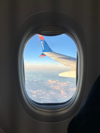 The image, taken from an airplane window, shows a view of the sky above Ambeli, Greece. The airplane's wing and tail fin are visible, with the tail fin featuring the "SunExpress" logo. Below the wing, a layer of clouds stretches across the horizon, tinged with the pink and orange hues of sunrise or sunset. The sky is a clear blue, and the airplane appears to be in flight. The interior of the airplane's cabin is also visible, with the dark interior of the cabin frame around the window. The overall scene conveys a sense of travel and aerial perspective.