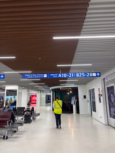 An interior shot captures a modern airport terminal, likely located in the city of Peschiera Borromeo, Italy, given the signage context. The scene is dominated by a high ceiling featuring a distinctive architectural design of alternating brown and white slatted panels. Fluorescent lighting is integrated into the ceiling, illuminating the space.

Below, the terminal floor is a polished, light-colored material, reflecting the overhead lights. Several rows of dark-colored airport seating are arranged in the foreground, some occupied by travelers. A man in a white t-shirt is seated, looking at his phone and resting his arm on a black suitcase. To his left, a woman and a man are seated, the man wearing glasses and a denim jacket.

In the mid-ground, a person wearing a bright yellow safety vest with "CUSTOMER SERVICE" text on the back walks away from the camera, heading towards a cafe labeled "VvTA Caffè." Behind the cafe, a red advertising display is visible.

Above the seating area, prominent blue directional signs provide information. One sign reads "Check-in lista d'attesa / Check-in waiting list" with a passenger icon. Another sign indicates "Imbarchi Gates A10-21 | B25-28" with a plane icon and an upward-pointing arrow. Further down the hallway, more signs and displays are present, including a fashion advertisement featuring a woman's face. The overall atmosphere is one of a clean, functional, and contemporary transportation hub.