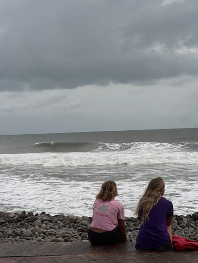 In El Tunco, El Salvador, two young women sit facing the ocean on a wooden deck, which is bordered by a collection of smooth, grey pebbles. The sky is overcast with heavy, dark clouds, suggesting impending rain or a storm. The ocean is turbulent, with large waves crashing ashore, creating white foam. The woman on the left wears a pink t-shirt with a graphic and the "Billabong" logo, and dark shorts. The woman on the right wears a purple t-shirt and dark shorts. A red bag rests on the deck to the right of the women.