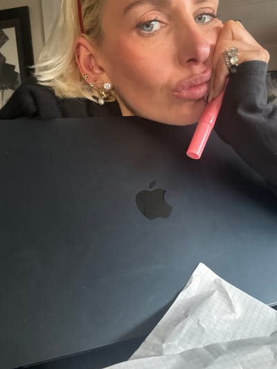 A close-up, rotated image shows a woman posing indoors, seemingly taking a selfie. Her face, seen from the nose upwards, features light-colored eyes with visible mascara and eyeliner, and blonde hair. Her lips are slightly puckered in a posed expression. Her visible ear is adorned with multiple intricate piercings, including studs and hoops.
Her hand, decorated with several rings, holds a pink tube, identified as a Clarins Lip Comfort Oil with "CLARIN*S" and "OIL" visible on its side, positioned near her mouth. The woman is positioned above a black laptop, clearly showing the Apple logo. To the right, a crinkled piece of white paper with grid lines is visible on the surface. A portion of a framed picture or artwork is partially visible in the lower background. The scene is set indoors, within the location context of Strömstad, Sweden.