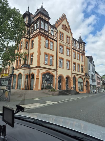 This image depicts a street scene in Bernkastel-Kues, Germany, specifically facing the Hotel Gassen at Cusanusstraße 7. The perspective is from inside a vehicle, looking forward and slightly to the right.
The most prominent structure is a large, multi-story building on the left, identified as "HOTEL-GASSEN" by lettering on its facade. This building features a light yellow-cream primary color with pronounced orange-brown architectural trim around windows, corners, and as decorative elements. The architecture includes traditional half-timbered sections on upper floors and gables, with dark grey slate roofs topped by multiple ornate spires and turrets. The ground floor of this building contains several arched storefronts and entrances; one prominent shop, "hees MODE," is visible on the far left with display windows showing mannequins. Steps lead up to several of the entrances from the sidewalk.
To the right of the Hotel Gassen, a row of interconnected buildings extends down the street. These structures exhibit varying pastel colors, including light blue, pink, and grey, and also feature traditional European architectural styles, some with half-timbering and decorative elements. Further down the street, two indistinct individuals are walking on the sidewalk away from the viewer.
The street in the foreground is asphalt, with a clear curb separating it from the sidewalk. White rectangular markings of a crosswalk are visible on the pavement. Reflections are present on the dashboard and lower windshield of the vehicle from which the photo was taken. A black navigation device is mounted on the dashboard in the lower-left corner. On the far left, partially obscured by a tree, a metal fence and a signpost are visible, with one sign mentioning "Brückenkeller."
The sky is partly cloudy, with significant grey cloud cover but also clear patches of blue sky visible, particularly towards the upper right. A tree with green foliage partially obscures the upper left portion of the main hotel building. In the distant background to the right, a vegetated hillside is visible, indicative of a river valley landscape.