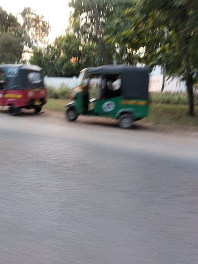The image depicts a street scene in Gombato Bongwe ward, Kenya. Two auto-rickshaws are positioned on the right side of a grey asphalt road, which occupies the lower half of the frame. In the midground, a green and yellow auto-rickshaw with a black canopy is visible, facing left, featuring a circular white emblem on its side. A blurred human figure is discernible inside its cab. Behind and to the left of the green vehicle, a red auto-rickshaw with a black canopy is partially visible, oriented with its rear towards the viewer. Both vehicles exhibit motion blur. Beyond the road, a grassy verge leads to a light-colored wall or fence, with dense green trees forming the background under ambient daylight conditions.