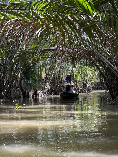 The image depicts a waterway in Tân Thạch, Vietnam, bordered by dense vegetation. A small boat occupies the center of the frame, carrying two individuals. One person, wearing a conical hat and a blue checkered shirt, is visible towards the front of the boat, possibly steering. The water reflects the surrounding vegetation and the sunlight, creating a shimmering effect. The environment is characterized by a tropical, lush landscape.