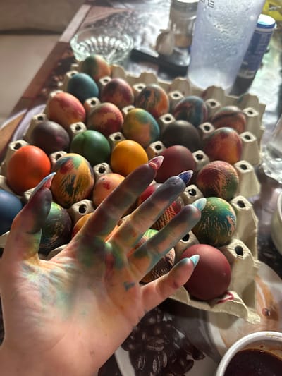 A close-up shot depicts a hand stained with dye resting above a carton of dyed Easter eggs. The hand has artificial nails with a light blue base, smudged with various colors of dye. The eggs display a range of colors and patterns, placed in individual compartments of the carton. A white bowl containing a dark liquid sits near the edge of the frame. Various containers are visible in the blurred background. The scene is set on a surface with a patterned design, possibly a countertop. The location in the city of Galiche, Bulgaria, cannot be determined from the given image.