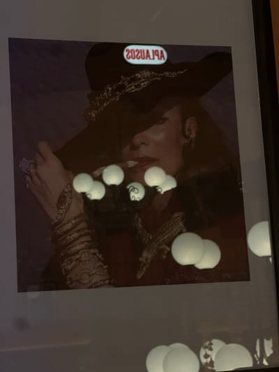 A framed, dark-toned portrait of a woman is depicted, viewed at an angle in an interior setting in Guanajuato City, Mexico. The woman, positioned centrally, wears a large-brimmed, dark hat with metallic or ornate trim, casting a shadow over her upper face. Her visible features include red-painted lips and an earring on her right ear. A cigarette is present in her mouth. Her left hand, adorned with multiple rings and a sleeve featuring intricate, possibly embroidered, patterns or stacked bracelets, is raised towards her face. Her attire also includes a distinct, small reptilian figure, resembling an alligator, positioned on the lower right side of her garment. The glass surface of the framed artwork exhibits reflections: numerous glowing, spherical lights, indicative of decorative string lights, and an inverted red sign that reads "APLAUSOS" (appearing as "SOZUAJLA" in reflection) positioned over the woman's hat. The environmental lighting is subdued, highlighting the reflections on the artwork.