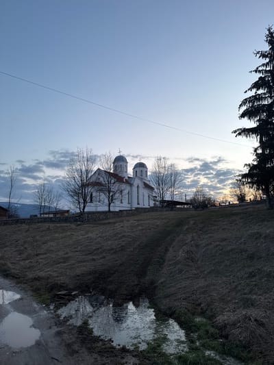 The image presents an exterior scene in Relyovo, Bulgaria, featuring a white Orthodox church positioned on an elevated grassy slope under a sky displaying shades of blue, white, and a faint orange at the horizon, indicating either dawn or dusk. The church structure, painted white with a reddish-brown tiled roof, prominently displays two domed steeples, each capped with a cross. Rectangular windows are visible on its facade. The church property is enclosed by a low stone wall. Several bare deciduous trees are situated around the church, their branches devoid of leaves. To the right of the frame, a tall, dark evergreen tree stands prominently. The foreground consists of a dirt path or track featuring multiple puddles of standing water, which reflect parts of the sky and the church's lower structures. A narrow water-filled channel also runs through the foreground. The ground is covered with dry, sparse brown and green grass. In the far left background, sections of other buildings are visible, with distant hills outlined against the sky. A thin utility line traverses the upper sky horizontally. No people, animals, or vehicles are present in the scene.