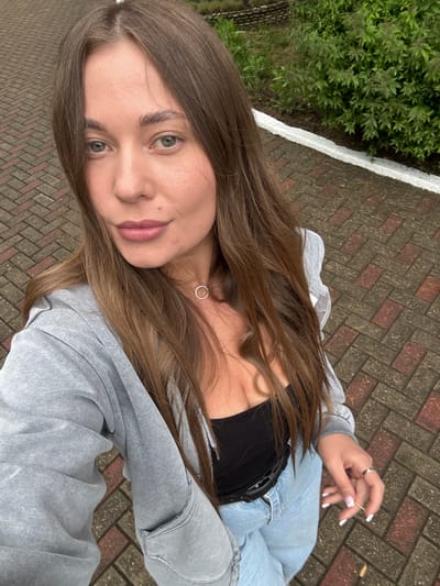 A young woman is pictured in a selfie. She has light brown hair, light eyes and fair skin. She is wearing a gray zip-up hoodie over a black top and light blue denim pants. She is wearing a silver necklace and a silver ring on her left hand, which is holding a thin stick. The background shows a brick walkway, green vegetation, and a white border. The location is potentially Sosnovy, Russia.