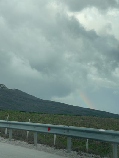 The image depicts an overcast sky with gray clouds and a faint rainbow appearing near a hillside. The hillside is covered in vegetation. In the foreground is a gray metal guardrail, situated beside a road in Buhovtsi, Bulgaria. A red reflector is visible on the guardrail.