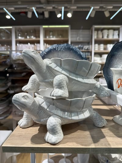 Two white ceramic turtle figurines are stacked on top of each other, placed on a light-colored wooden surface. The figurines are detailed with textured shells and heads. The background shows shelves filled with various items, seemingly in a store or display setting. Due to the lack of additional information, the specific location cannot be determined with certainty. The city of Murmansk, Russia cannot be confirmed as the location.