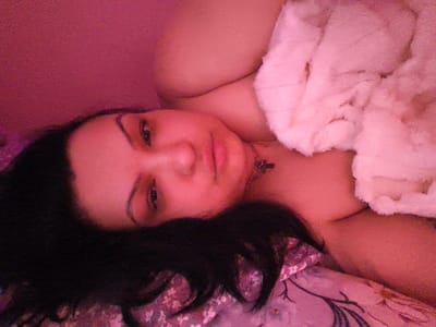 The image shows a close-up of a person's face and upper body, captured from a low angle. The individual has dark hair, brown eyes, and is wearing a necklace with a pendant. They are lying down, with a pinkish blanket or towel covering part of their body. The background is a solid pink color, suggesting an indoor setting with artificial lighting. There are no discernible elements in the image that would indicate a specific location within the city of Komotini, Greece. The image is grainy and has a soft focus, contributing to a relaxed or intimate atmosphere.