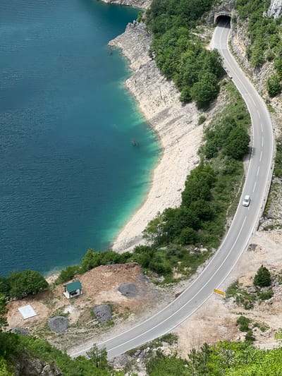 The image is an aerial view of a section of road and lake in Plužine, Montenegro. The road curves along the lake's edge, passing through a tunnel at one end. A white car is traveling along the road. The lake is a deep blue color with turquoise hues along the shore. The shoreline shows exposed rock and sparse vegetation. Along the road, there is a yellow road sign. Further along the shoreline, there is a small structure with a green roof and an adjacent white concrete slab. The surrounding terrain consists of rocky cliffs and green trees.