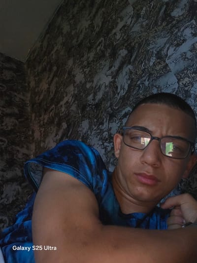 The image shows a male individual taking a selfie indoors, with the location described as Konyavo, Bulgaria. The individual, appearing to be a young adult, has short dark hair and wears dark-rimmed glasses that reflect a window with hints of green foliage outside. He has a neutral expression, looking slightly downward and to his right. He is wearing a short-sleeved shirt with a blue and black tie-dye or swirling pattern. His left arm is bent, with his hand resting near his chin or jawline. The background features a wall covered in a distinct dark, swirling, marble-like pattern composed of black, blue, gray, and subtle reddish-brown tones. A corner of a plain, lighter gray wall or ceiling is visible in the upper left quadrant. A watermark reading 'Galaxy S25 Ultra' is present in the lower left corner of the image.
